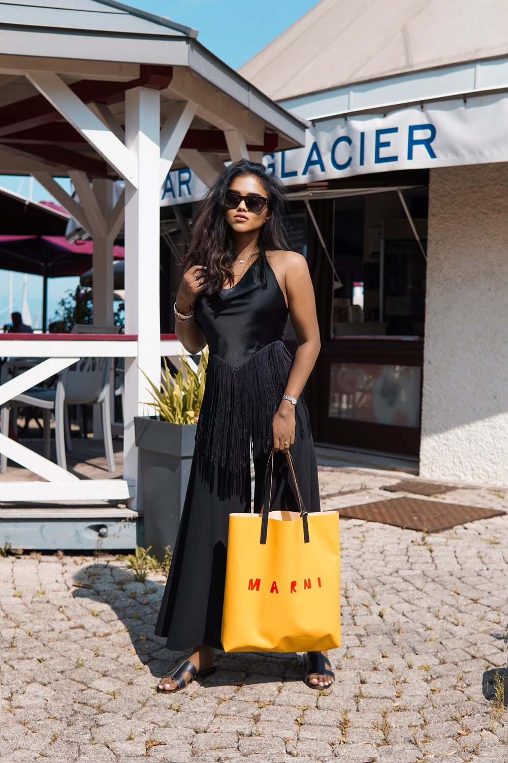 Sachini in front of a cafe wearing a black dresses and sunglasses holding a yellow Marni bag